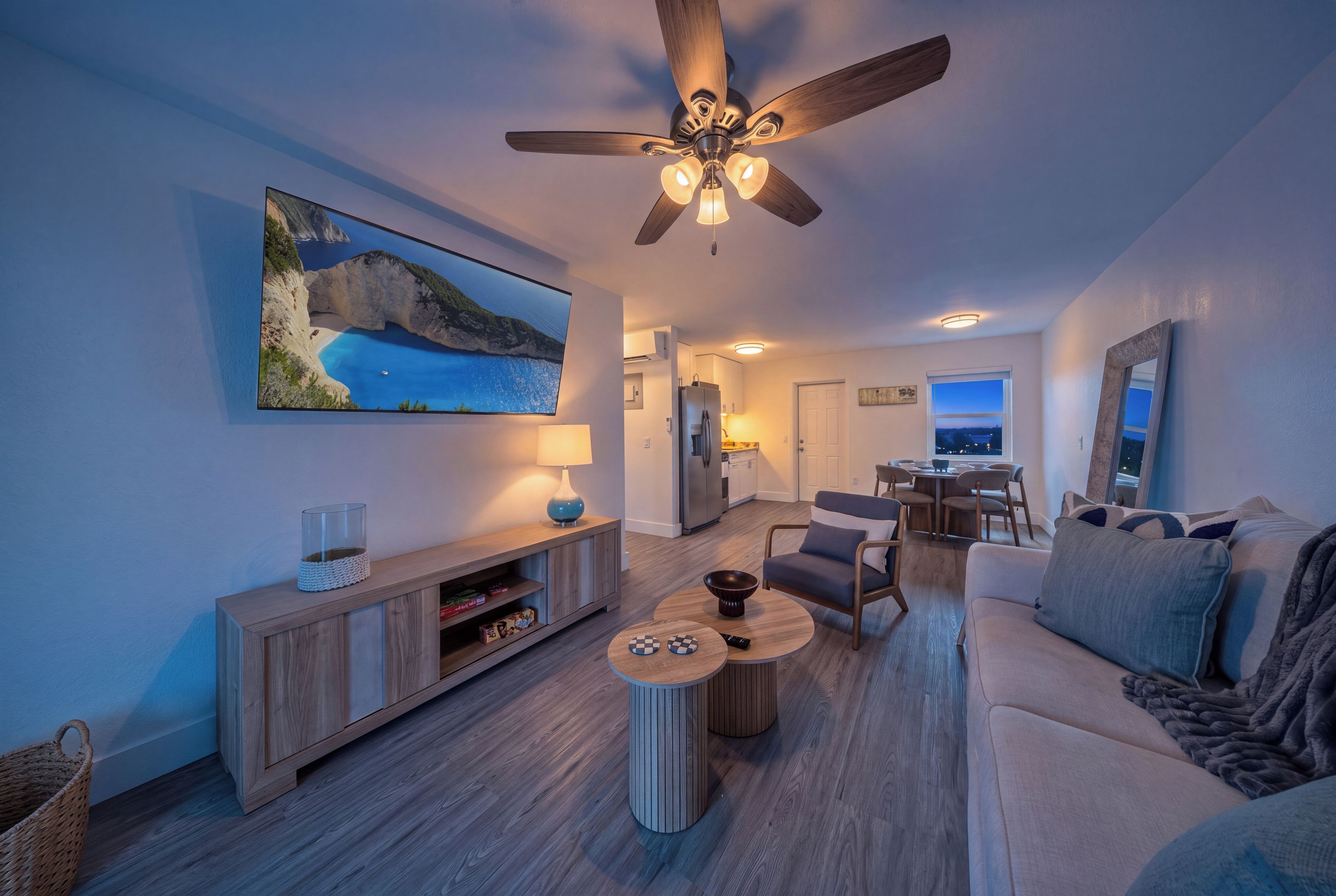 A modern living room with a ceiling fan, a flat-screen TV mounted on the wall, a dining area with a table and chairs, and a cozy seating area with a sofa.