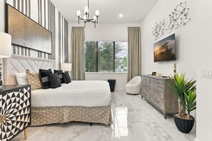A modern bedroom with a large bed, a bedside table, a dresser, a chair, a potted plant, and a window with blinds. - Luxury 15BR Suites, Pool and Themed Bedrooms (Williamsburg)