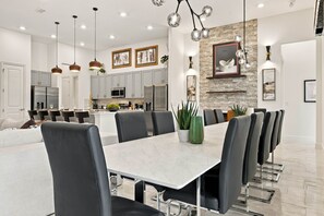 A modern dining area with a marble table and black chairs, a kitchen in the background, and pendant lighting. - Luxury 15BR Suites, Pool and Themed Bedrooms (Williamsburg)