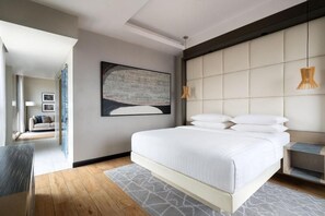Una habitación de hotel moderna con una cama grande, un cabecero, un cuadro en la pared y vistas al área de estar contigua.