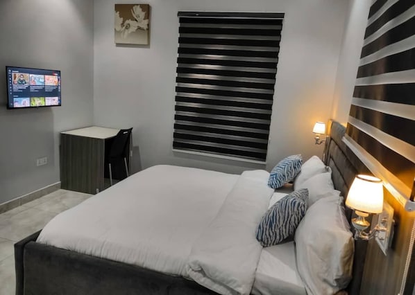 A bedroom with a bed, a TV mounted on the wall, a bedside lamp, and a window with blinds.