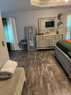 A bedroom with a wooden floor, a bed with a green blanket, a dresser with drawers, a large mirror, and a window with curtains. - Cozy Family Home away from Home
3-bedroom house in beautiful Pooler w/everything (Pooler)