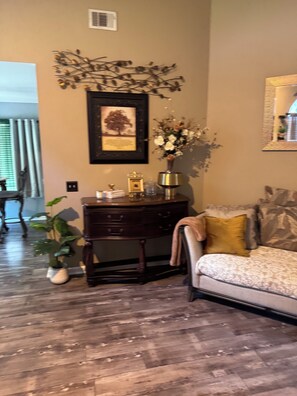 A room with a wooden floor, a desk with drawers, a vase with flowers, a framed picture on the wall, and a couch with a cushion. - Cozy Family Home away from Home
3-bedroom house in beautiful Pooler w/everything (Pooler)