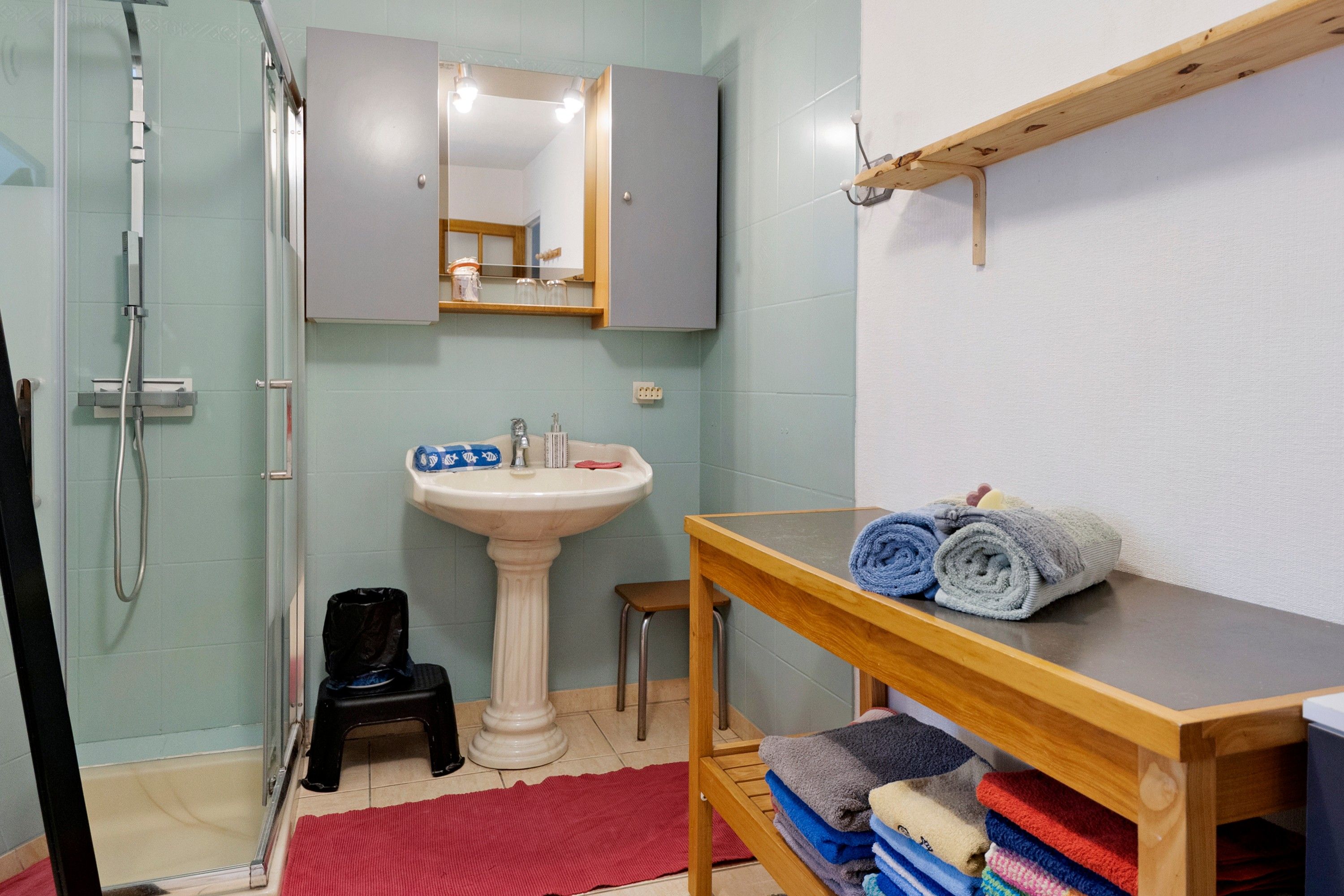 A bathroom with a shower, sink, mirror, and a wooden shelf with towels.
