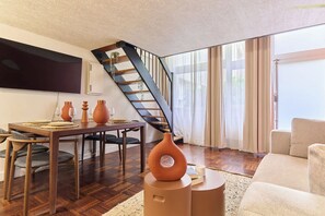A modern living room with a wooden dining table set for four, a sofa, a coffee table with a decorative vase, and a staircase with a geometric patterned ceiling. - Apartment Le Marais/ Place des Vosges (Paris)