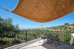 A balcony with a table and chairs, a thatched umbrella, and a view of a hilly landscape. - Apartment for two among olive groves in Andalusia (Alcaucin)