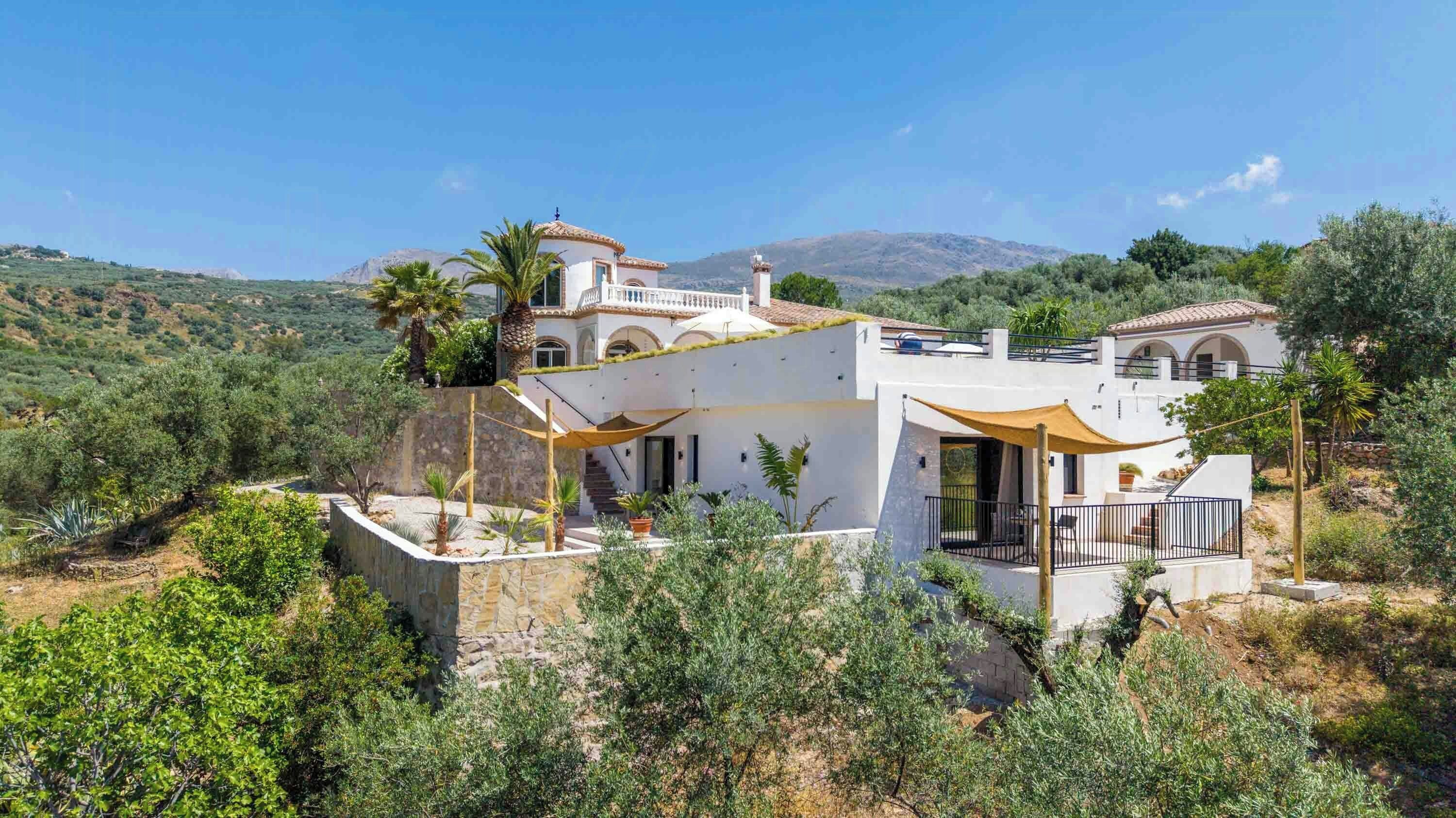 A modern villa with a stone retaining wall, a patio with outdoor furniture, and a view of the surrounding hills.