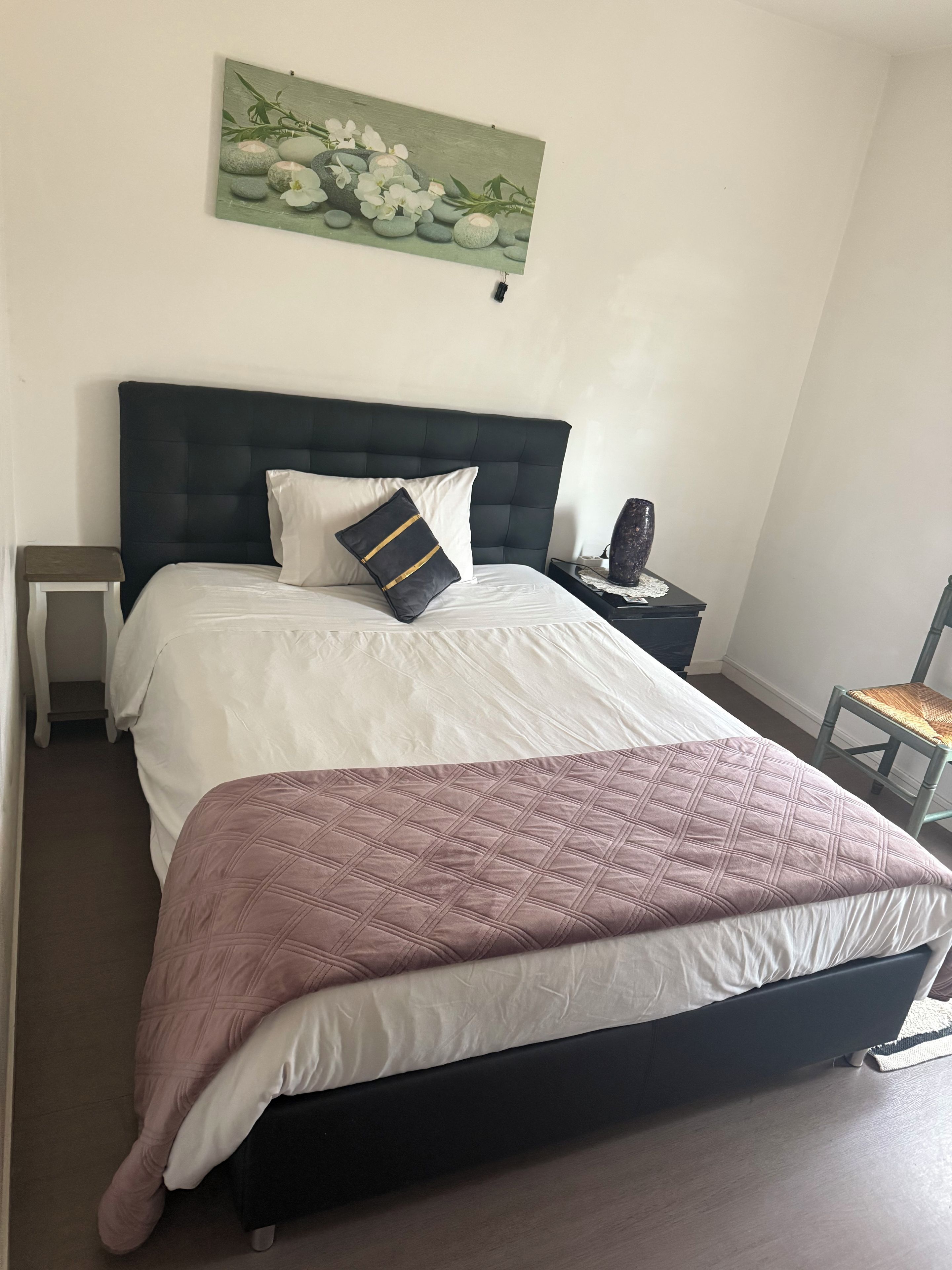 Een slaapkamer met een bed, een nachtkastje, een stoel en een schilderij aan de muur.