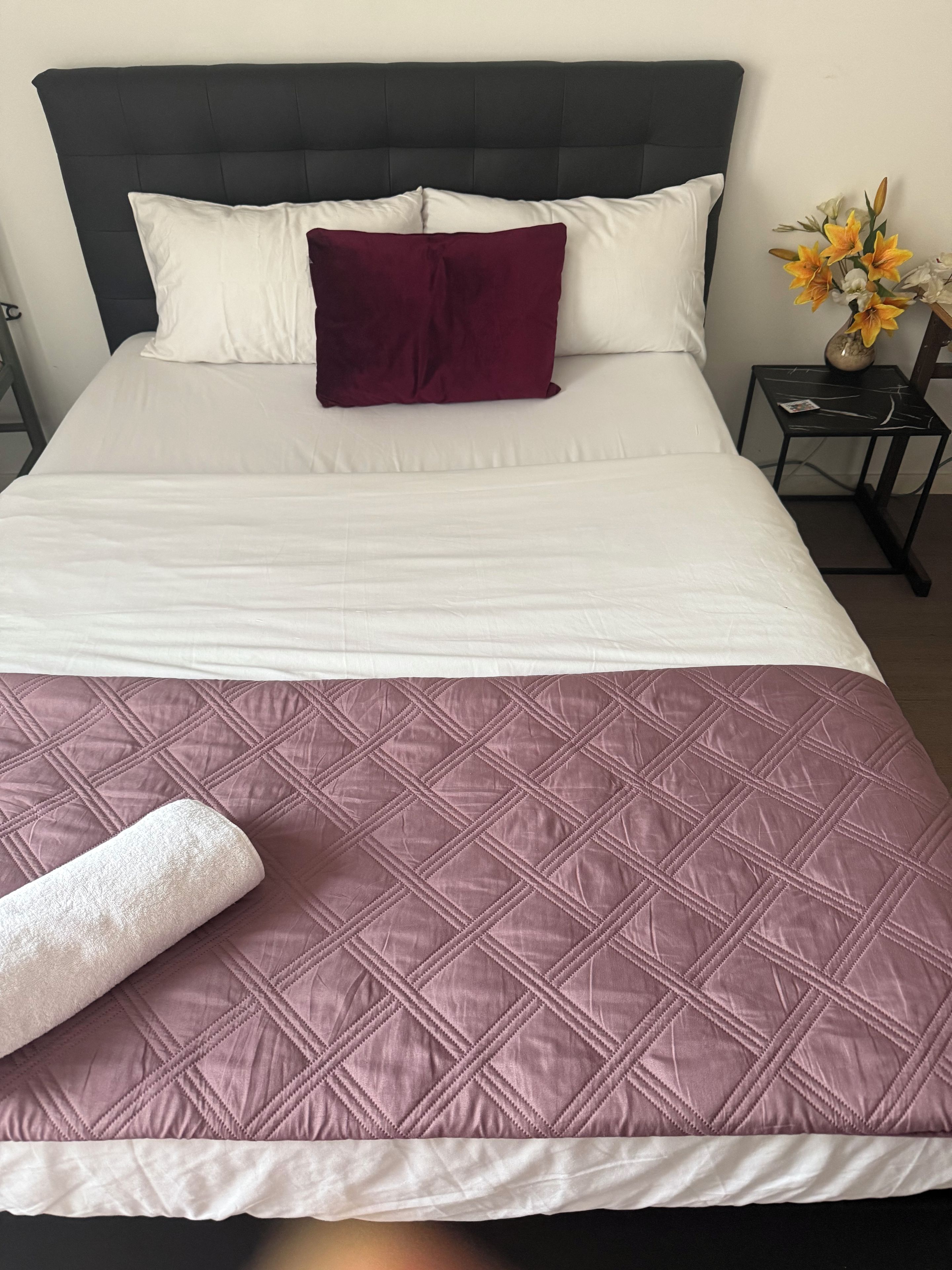 Een bed met een donker hoofdbord, witte en bordeauxrode kussens, een paars gewatteerd sprei en een witte handdoek op het bed.