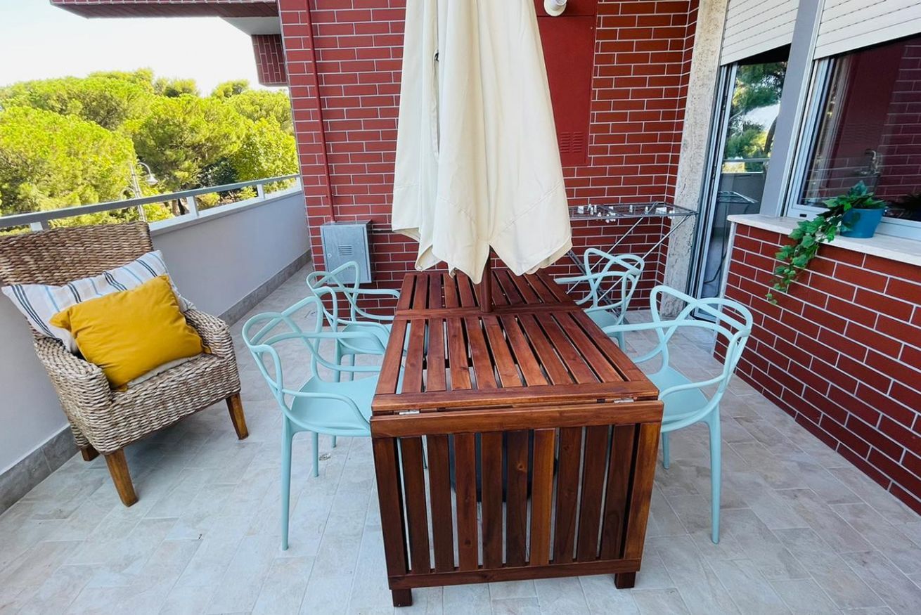 Un balcone con un tavolo e sedie in legno, una sedia in vimini con un cuscino giallo e una pianta in vaso.