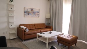 A living room with a brown leather sofa, a white coffee table, and a small ottoman. There is a painting on the wall and a floor lamp. - Walk to Beach Modern 1-Bed Apt CasaAmarilla205 (Oranjestad)