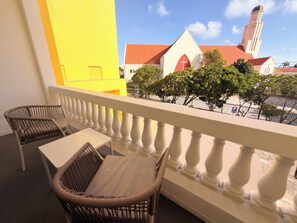 A balcony with wicker furniture and a view of a building with a red roof and a tower. - Walk to Beach Modern 1-Bed Apt CasaAmarilla205 (Oranjestad)