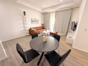 Ein modernes Wohnzimmer mit einem runden Esstisch, schwarzen Stühlen, einem braunen Ledersofa, einem weißen Couchtisch und einem an der Wand befestigten Fernseher.