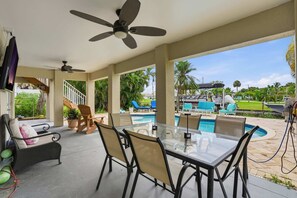 A covered outdoor area with a glass table and chairs, a ceiling fan, and a view of a swimming pool and palm trees. - Pet-Friendly Canal Retreat  Pool & Balconies (Bonita Springs)