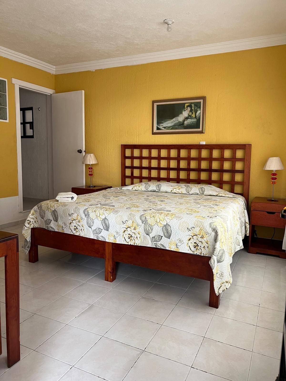 A bedroom with a wooden bed, a bedside table, a nightstand, a door, and a painting on the wall.