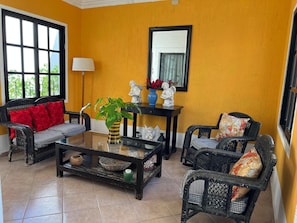 A room with a yellow wall, a black-framed mirror, wicker furniture, and a glass coffee table with decorative items.