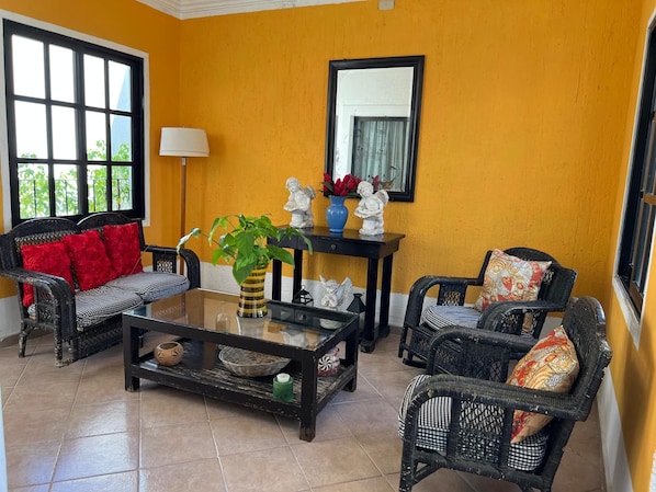A room with a yellow wall, a black-framed mirror, wicker furniture, and a glass coffee table with decorative items.