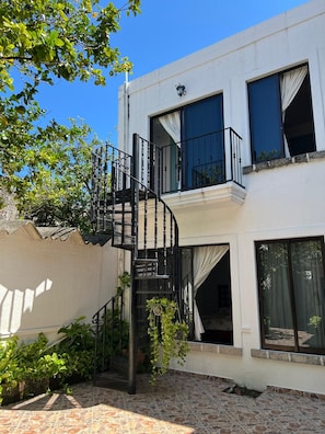 Un edificio de dos plantas con una escalera espiral que lleva a un balcón, rodeado de follaje y un patio con suelo de piedra.