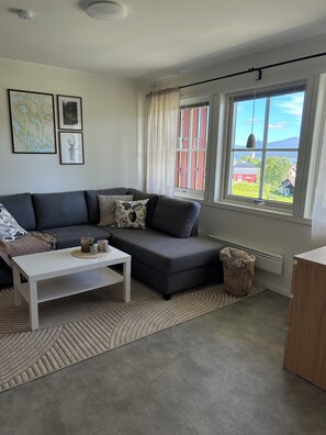Uma sala de estar moderna com um sofá cinzento, uma mesa de centro branca e uma janela grande com vista para um edifício vermelho e áreas verdes.