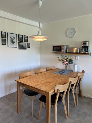 Uma sala de jantar com mesa e cadeiras de madeira, uma luminária pendente, um relógio e quadros nas paredes.