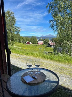 Mesa de vidro redonda com duas taças de vinho e dados, um cardápio, e vista para paisagem rural com casas e uma igreja.