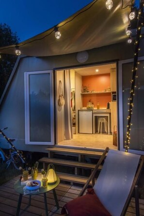 Un área de comedor al aire libre con una mesa y sillas, una pequeña cocina visible a través de una puerta abierta, y luces colgantes.