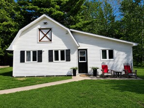 A white single-story house with a gabled roof, a front door, and a small porch with two chairs and a table. - White Pine Cottage- Family Friendly, Play Structure, Huge Yard and Serenity (Richibucto Road)