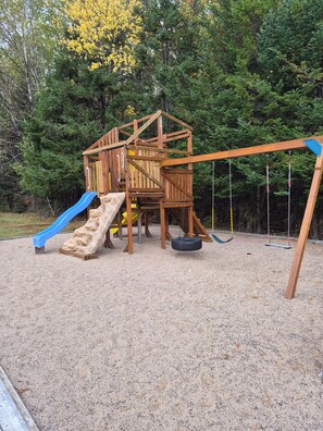 A wooden play structure with a slide and swings, set on a gravel surface with trees in the background.