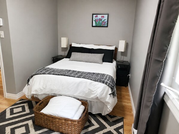 A neatly made bed with a white comforter and a grey throw, flanked by two bedside tables with lamps, a framed floral artwork on the wall, and a woven basket with a white pillow on the floor.