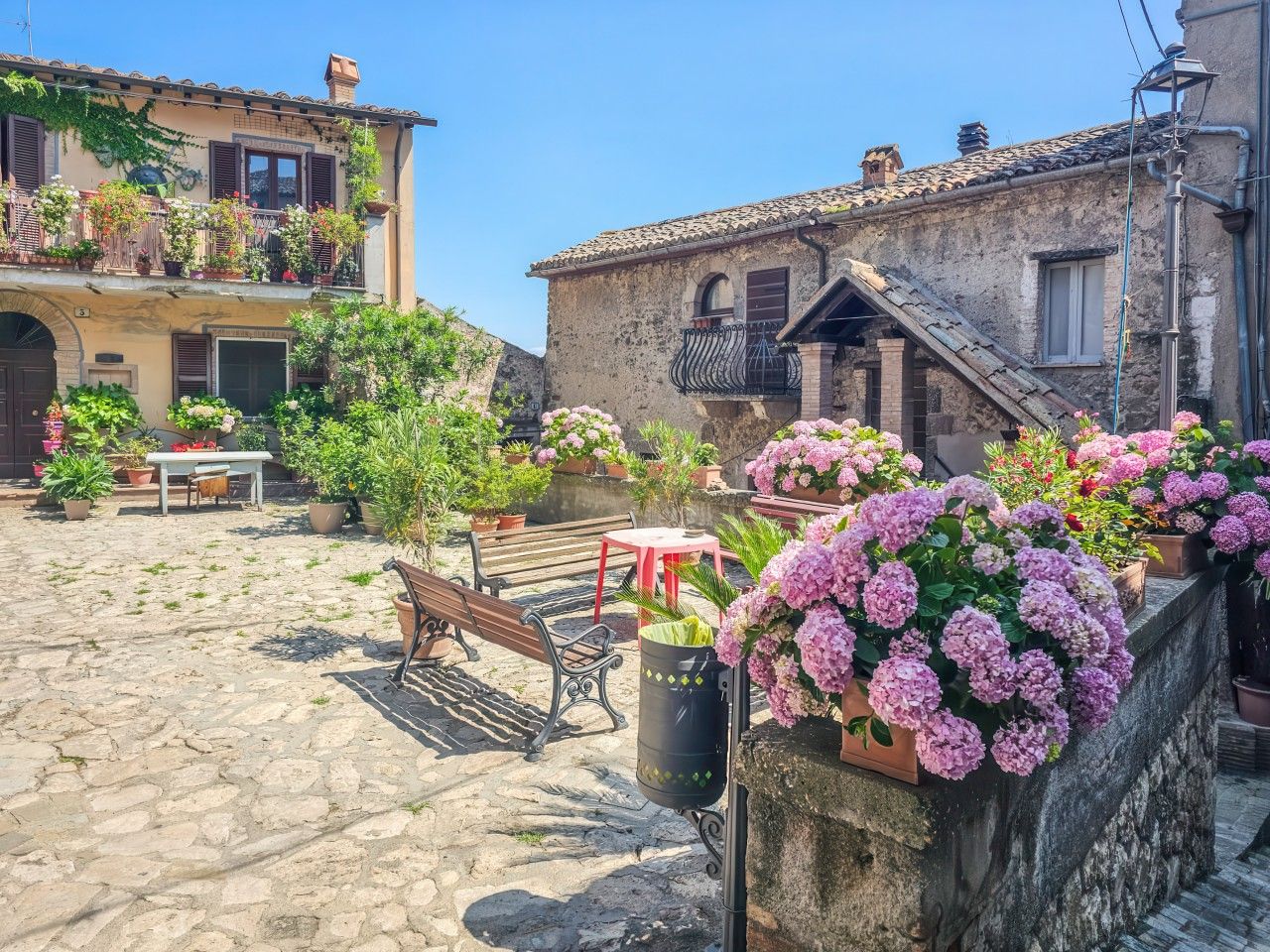 A cobblestone courtyard with benches, potted plants, and a building with a balcony.