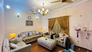 A living room with a sofa set, a glass coffee table, a chandelier, a ceiling fan, and a clock on the wall. - Feel Like Home in a Luxury, Secured and Peaceful 2 Story House (Karachi)