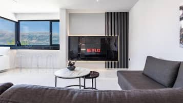 A modern living room with a large flat-screen TV, a sofa, a coffee table, and a view of the outdoors.