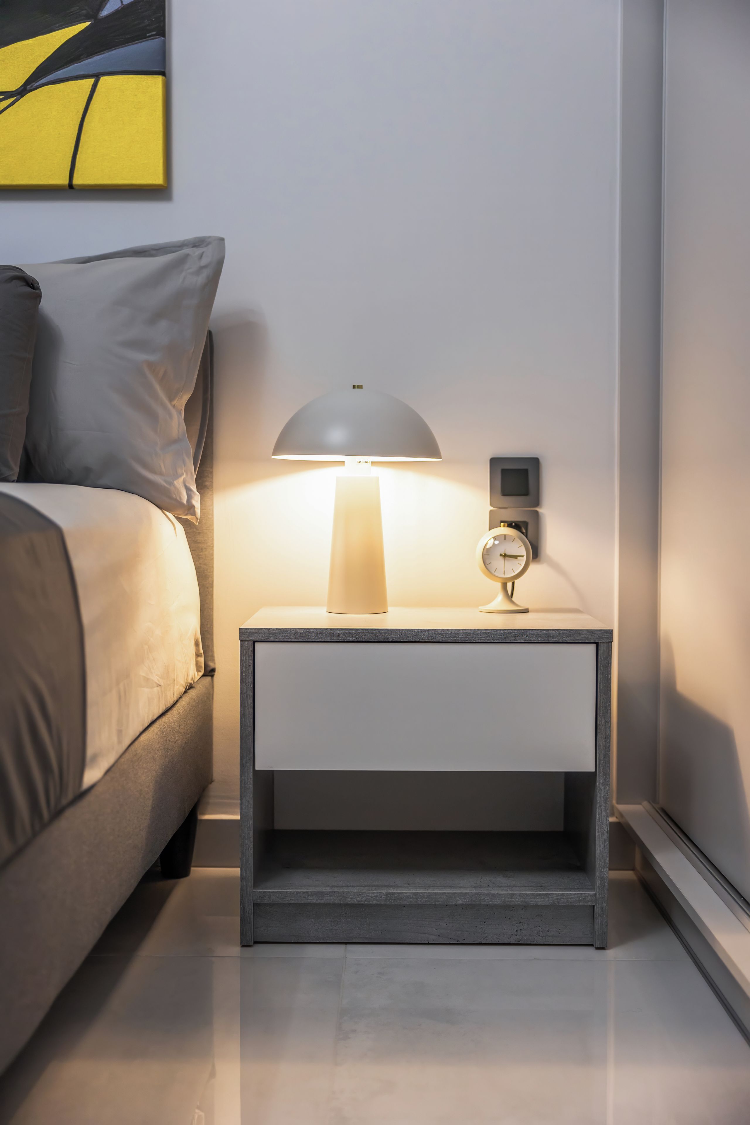 A modern bedroom with a bed, a bedside table with a lamp, and a wall-mounted clock.