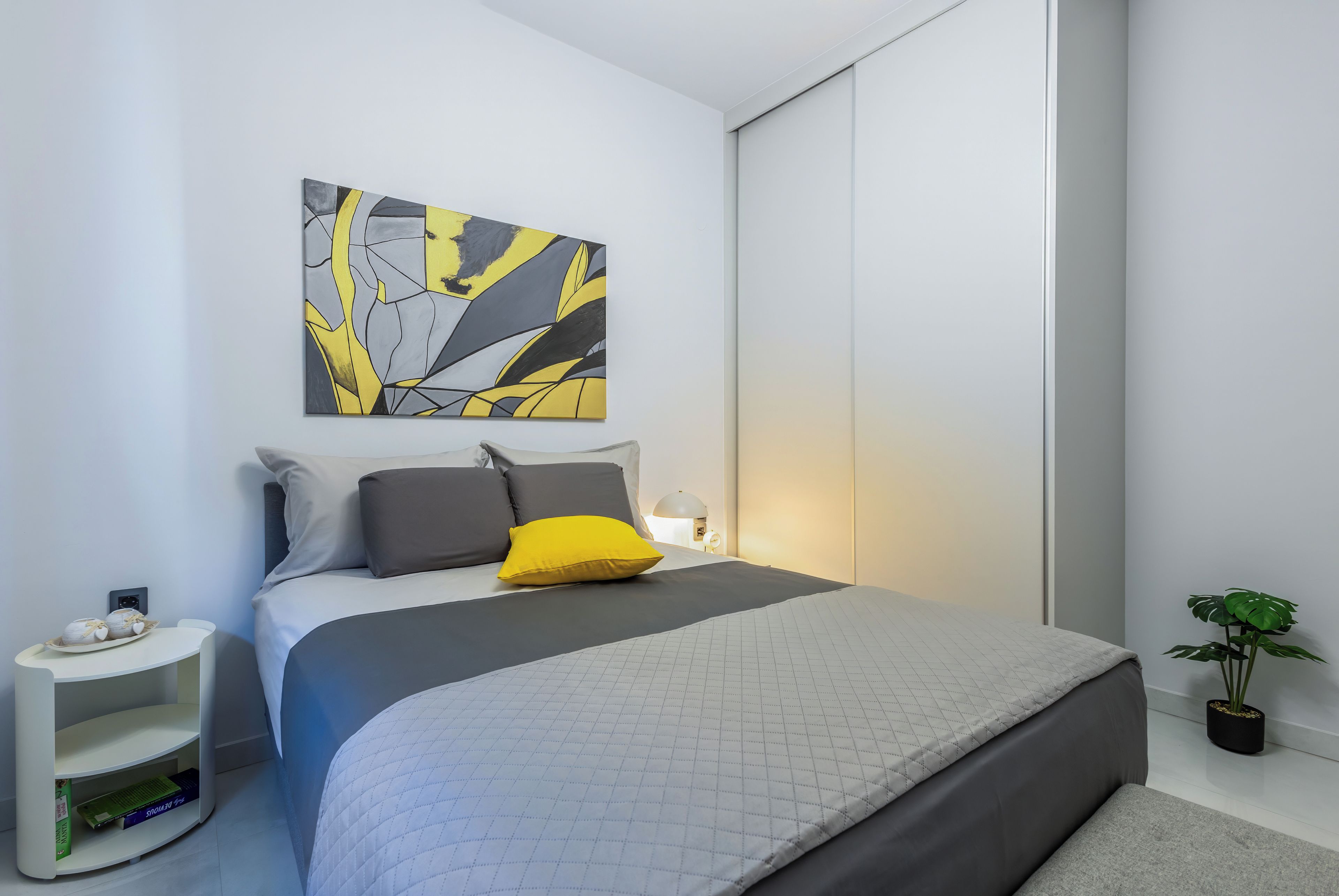Une chambre à coucher moderne avec un lit, des oreillers, une table de chevet, une œuvre d’art murale et une plante en pot.