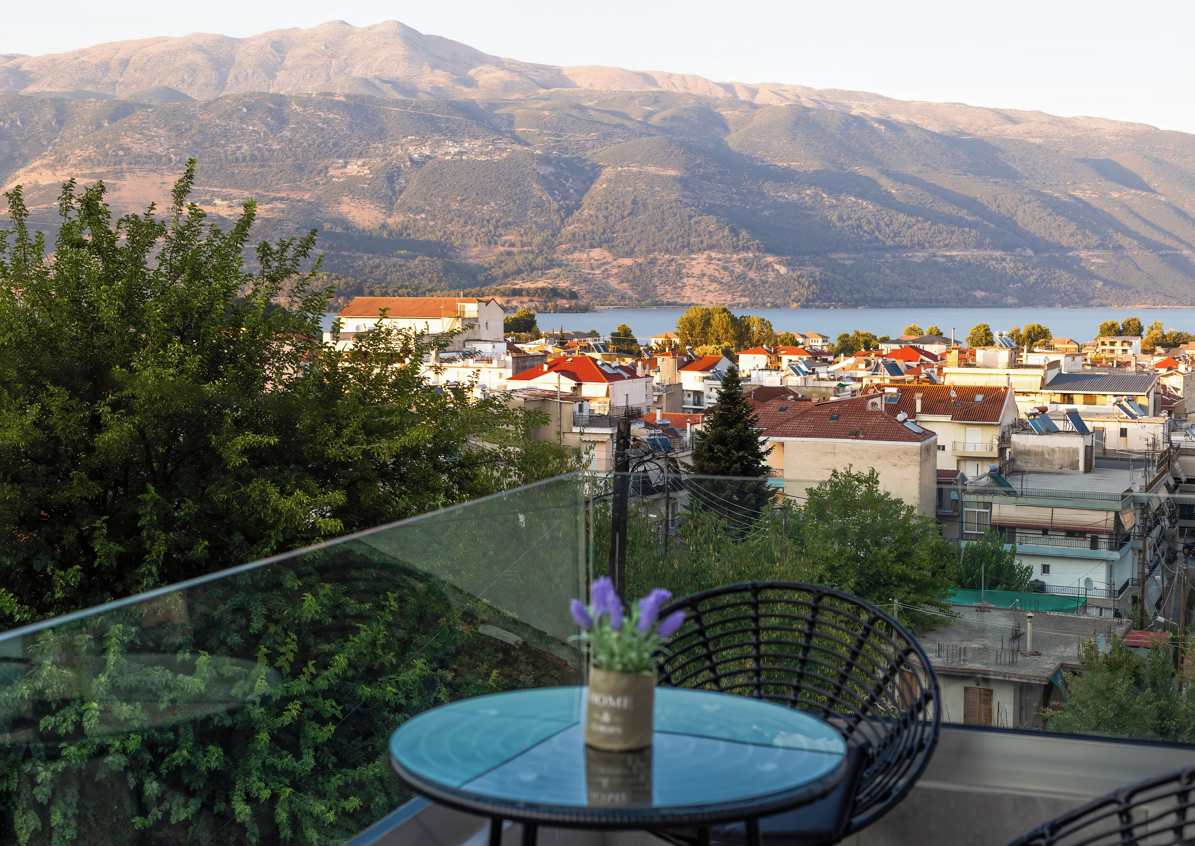 A balcony with a glass table and chairs, overlooking a town with buildings and mountains in the distance.