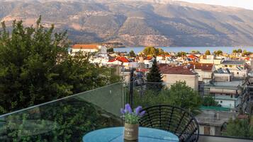 A balcony with a glass table and chairs, overlooking a town with buildings and mountains in the distance.