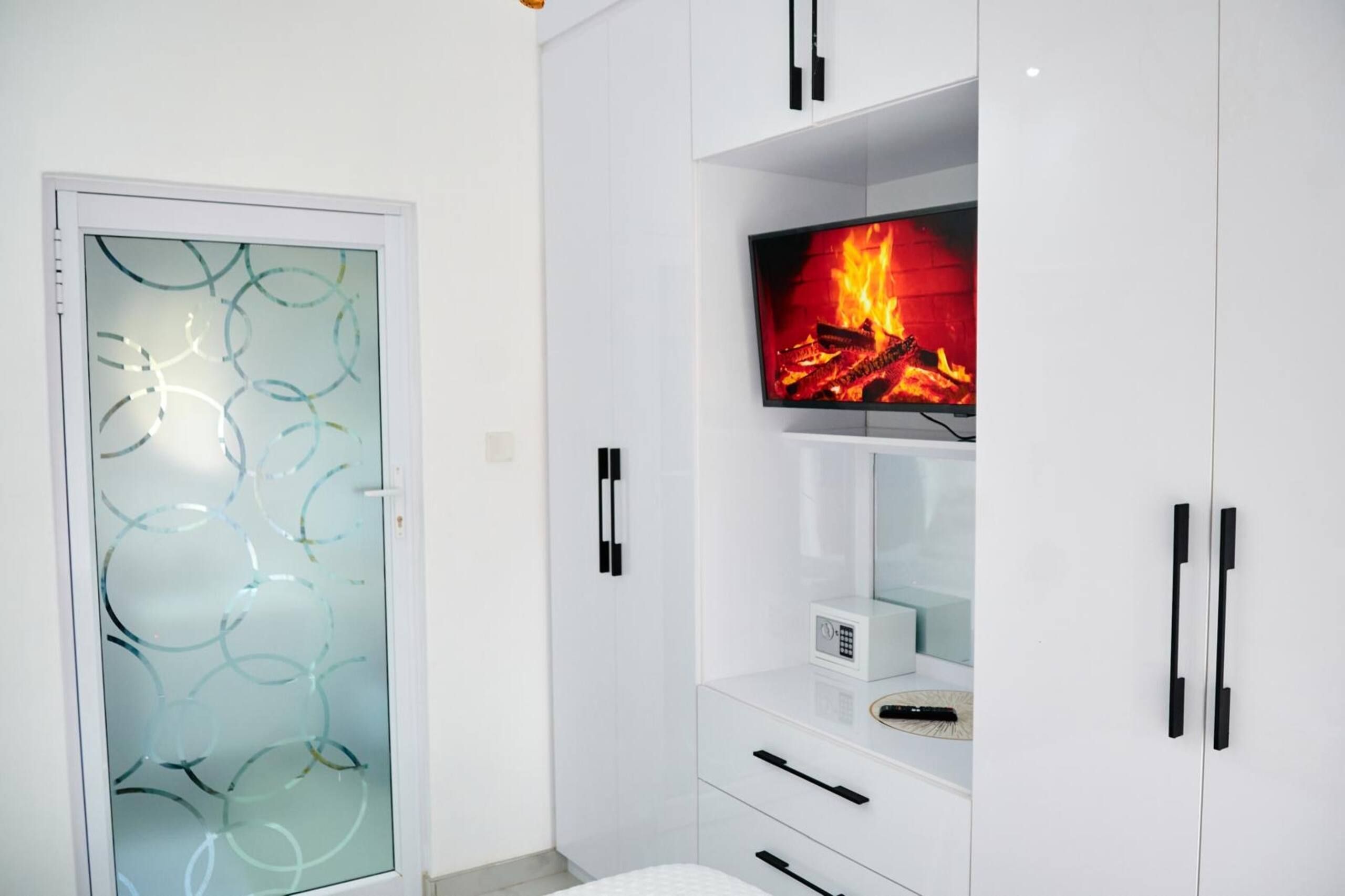 Un baño moderno con puerta de vidrio, muebles blancos y una televisión que muestra una chimenea.