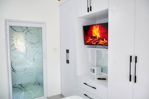 Une salle de bain moderne avec une porte en verre, des meubles blancs et un téléviseur diffusant une image de cheminée.