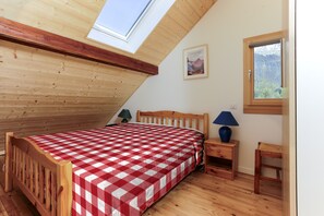 A bedroom with a sloped ceiling, a bed with a checkered bedspread, a nightstand with a lamp, a small table with a chair, and a window with a view of trees. - Jacques’s Treasure: A cosy, renovated chalet in the Chartreuse (Saint-Pierre-d'Entremont)