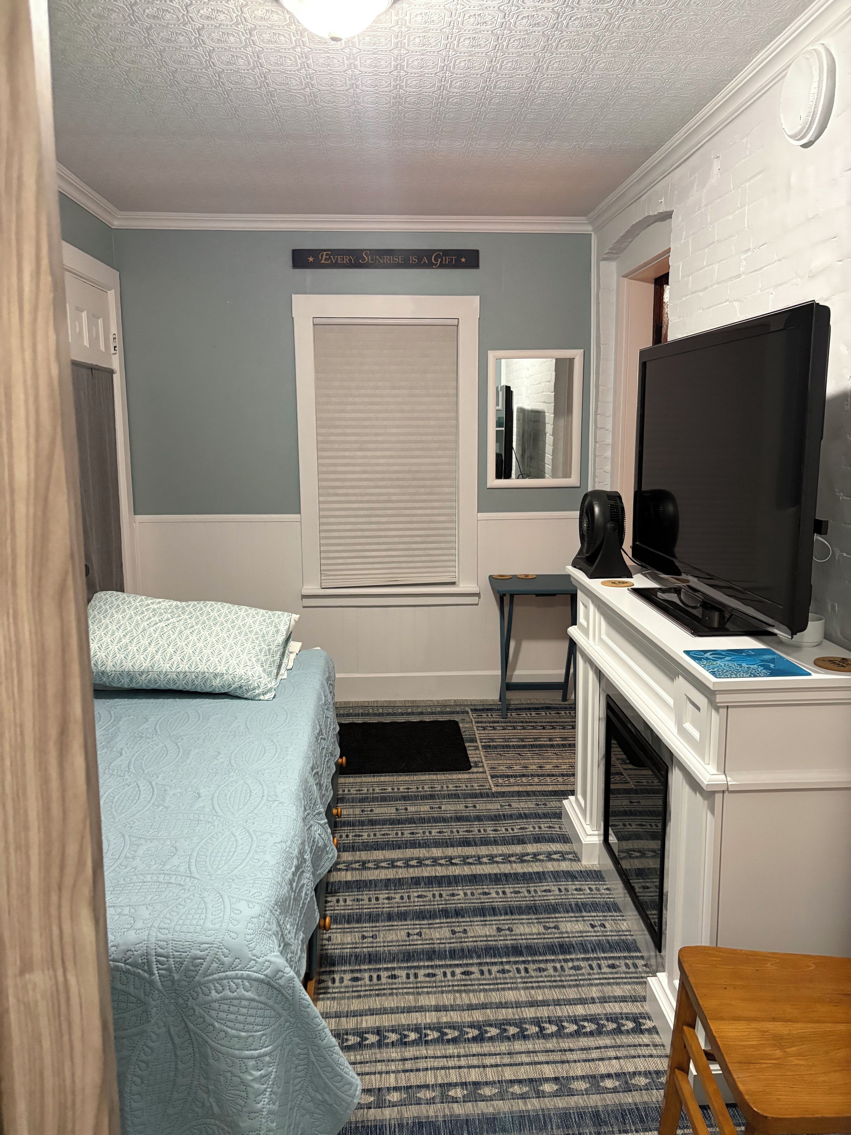 A room with a bed, a desk with a television, a window with a view, and a door.