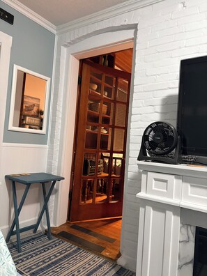 Una habitación con chimenea de ladrillo blanco, televisor, una mesita, alfombra estampada y una puerta de madera que da a otra habitación con estanterías.