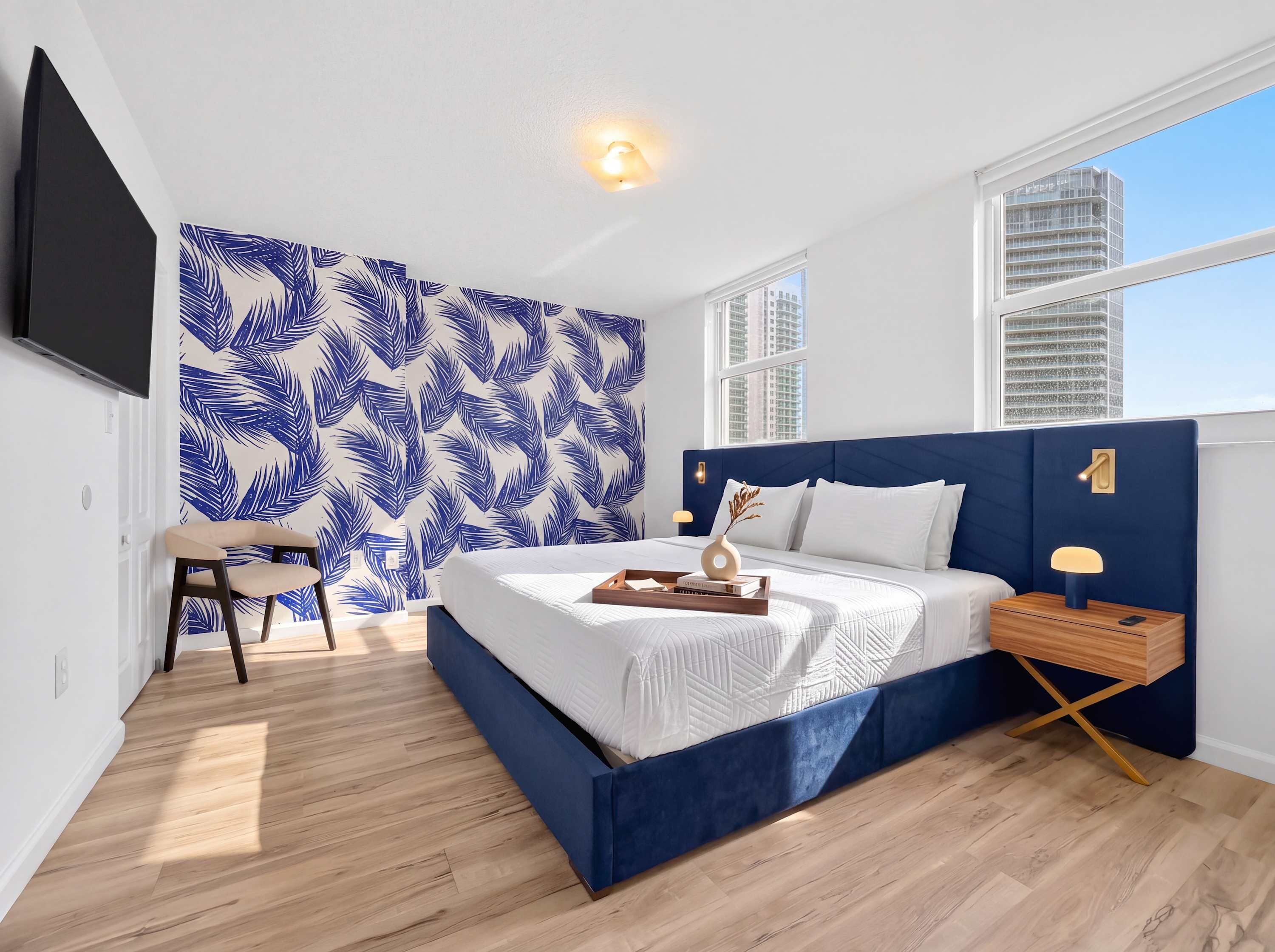 A modern bedroom with a large bed, a nightstand, a chair, a wall-mounted TV, and a window with a city view.