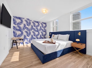 A modern bedroom with a large bed, a nightstand, a chair, a wall-mounted TV, and a window with a city view. - 2BR Brickell Condo With Pool, Gym & Balcony Views (Miami)
