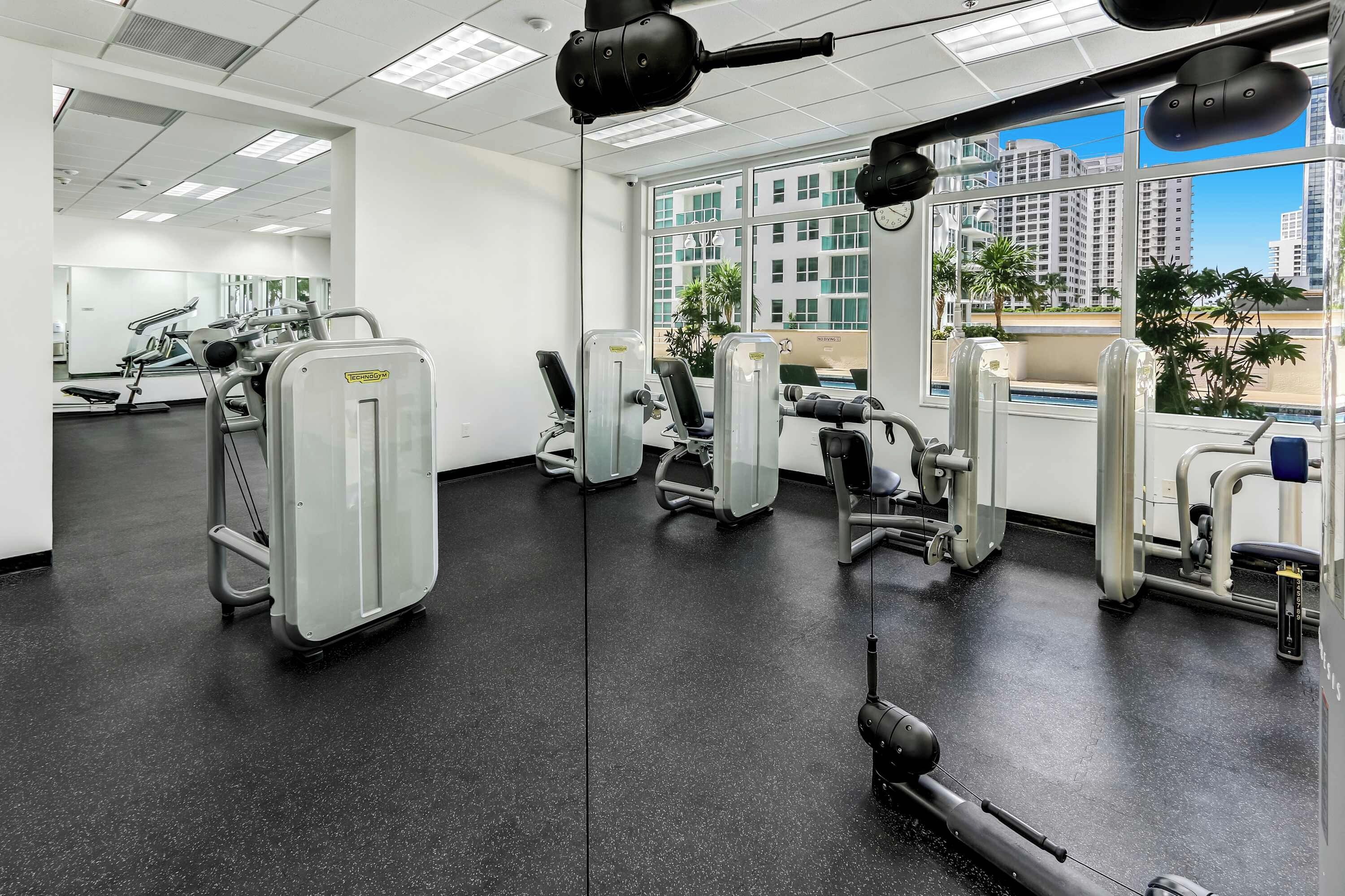 A gym with various exercise machines, large windows, and a view of buildings.