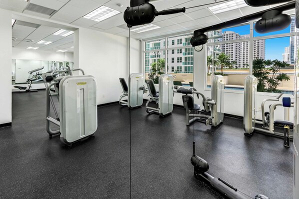 A gym with various exercise machines, large windows, and a view of buildings. - 2BR Brickell Condo With Pool, Gym & Balcony Views (Miami)