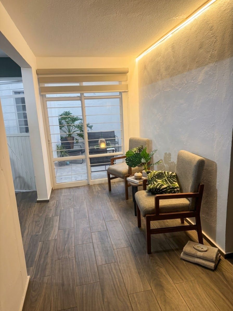 A room with a sliding glass door leading to an outdoor area, a wooden floor, two armchairs, a small table with a plant, and a wall-mounted light fixture.