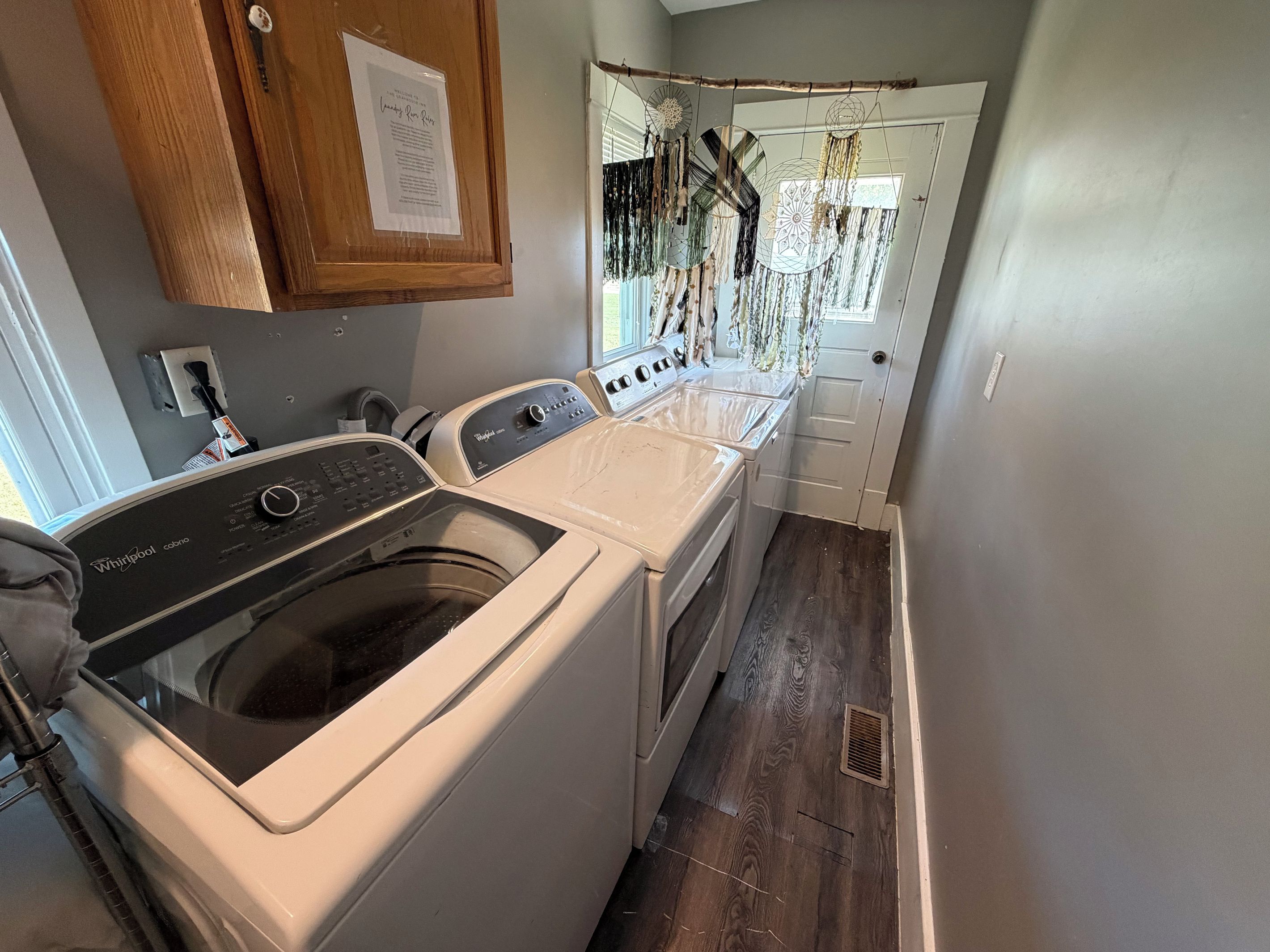 A laundry room with a Whirlpool washer and dryer, wooden cabinets, and a door with a hanging dreamcatcher.