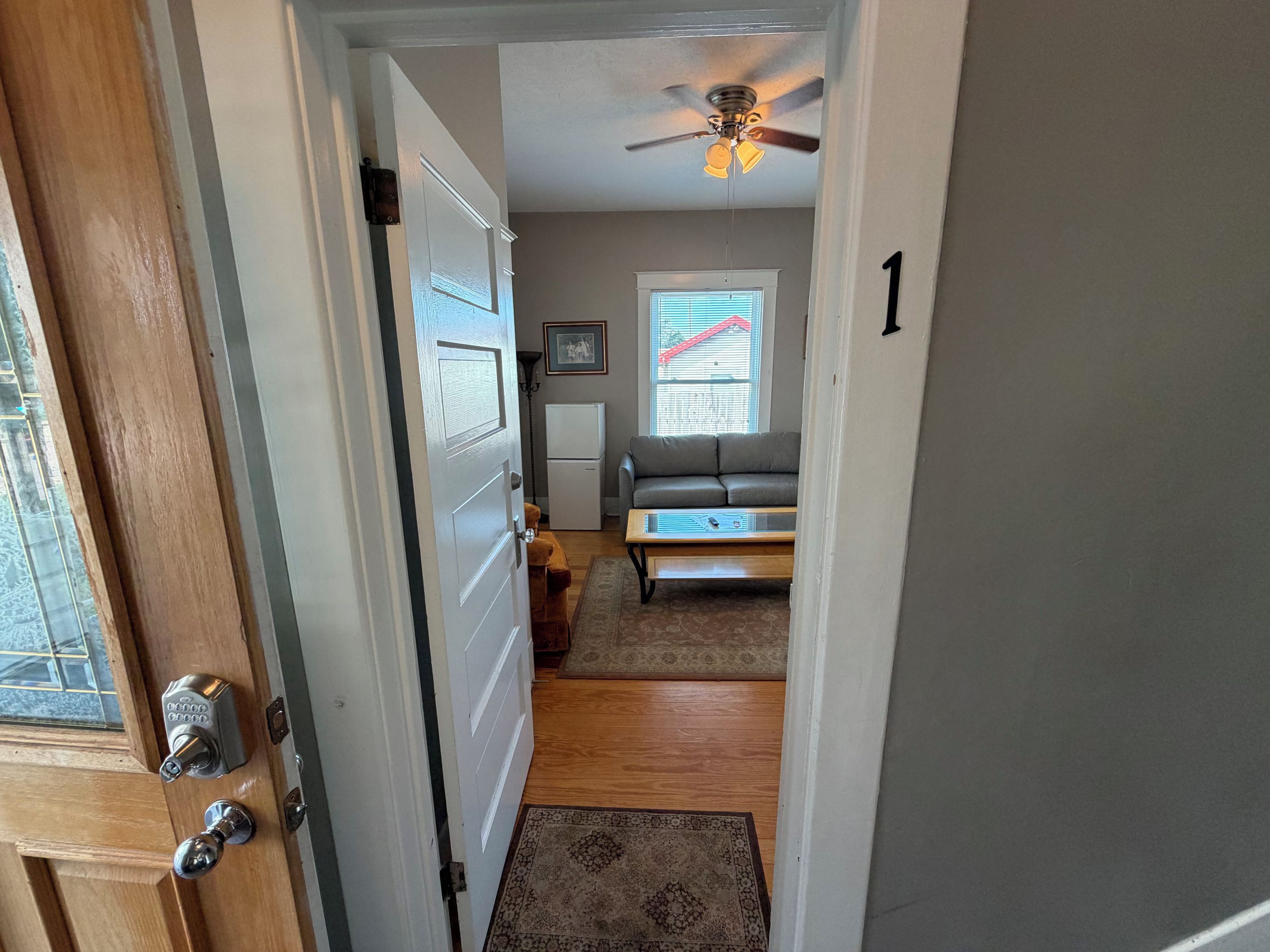 A hallway leading to a living room with a ceiling fan, a sofa, a coffee table, and a refrigerator.