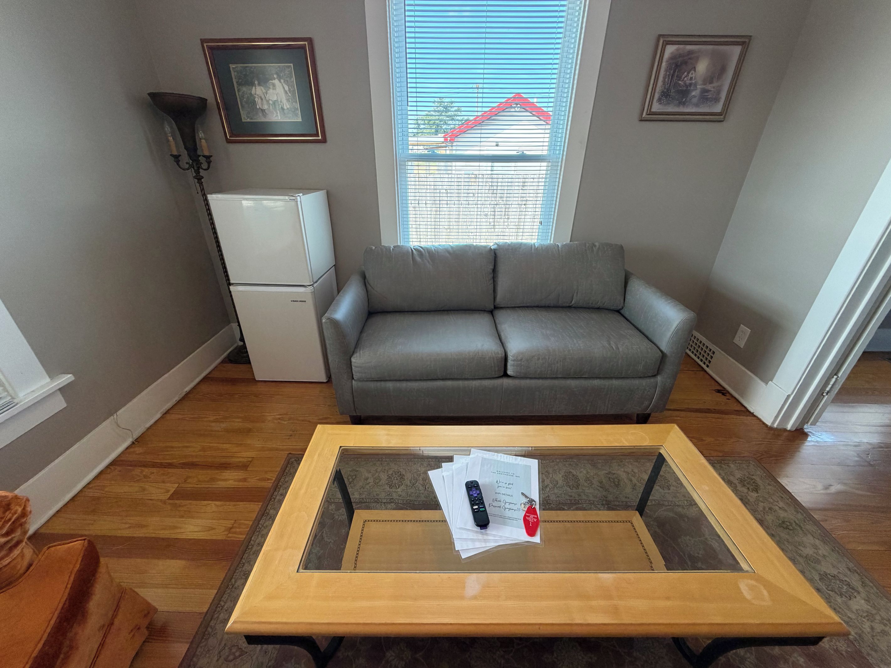 A living room with a grey sofa, a small white refrigerator, a glass-top coffee table with papers and a remote, and a window with blinds.