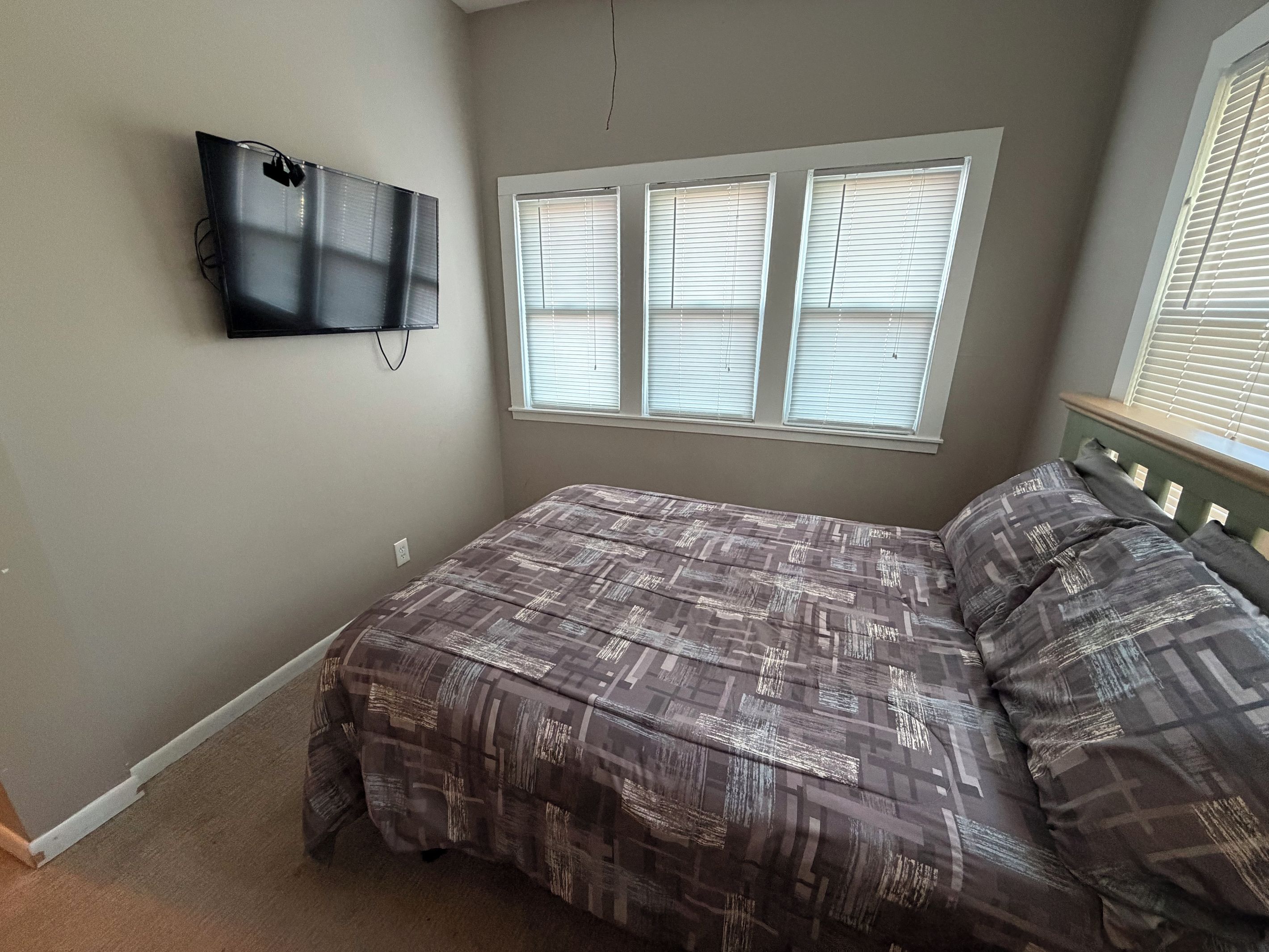 A bedroom with a bed, a television mounted on the wall, and a window with blinds.